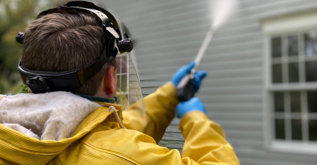 Person cleaning house siding with a pressure washer, wearing protective gear in a yellow raincoat.